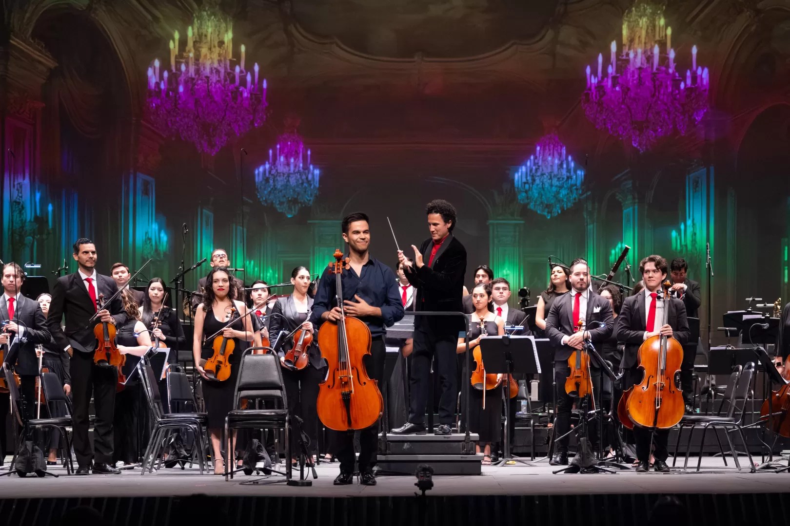 Ein Sinfonieorchester steht zum Applaus, ganz vorne in der Mitte steht lächelnd ein Cellist. Schräg hinter ihm applaudiert der Dirigent.
