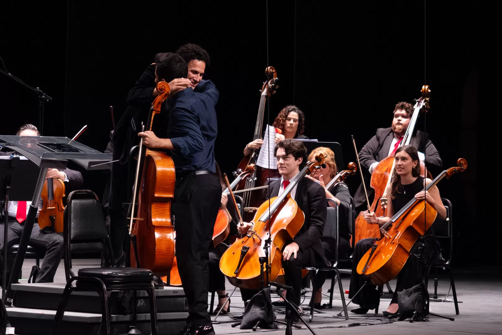Ein Dirigent und ein Cellist umarmen sich vor dem Orchester.