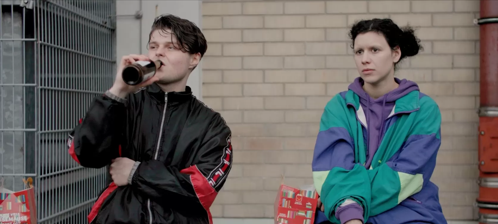 Zwei junge Personen in Jogginganzügen, einer trinkt ein Bier aus der Flasche.