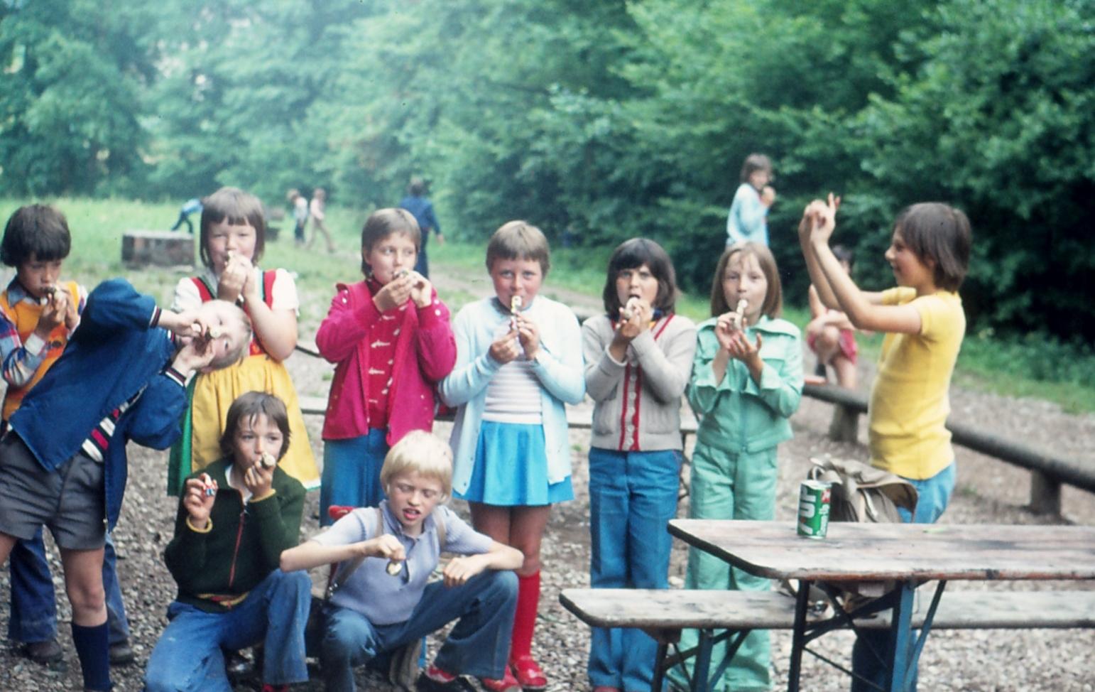 altes Foto. Gruppe von Kindern steht draußen und spielt Blockflöte, die junge Susanne Rohn steht rechts und dirigiert
