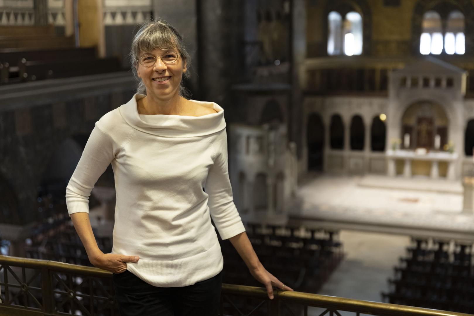 Susanne Rohn trägt ein schickes, weißes Oberteil und hält sich an der Emporenbrüstung fest. Blick in Kirchenraum