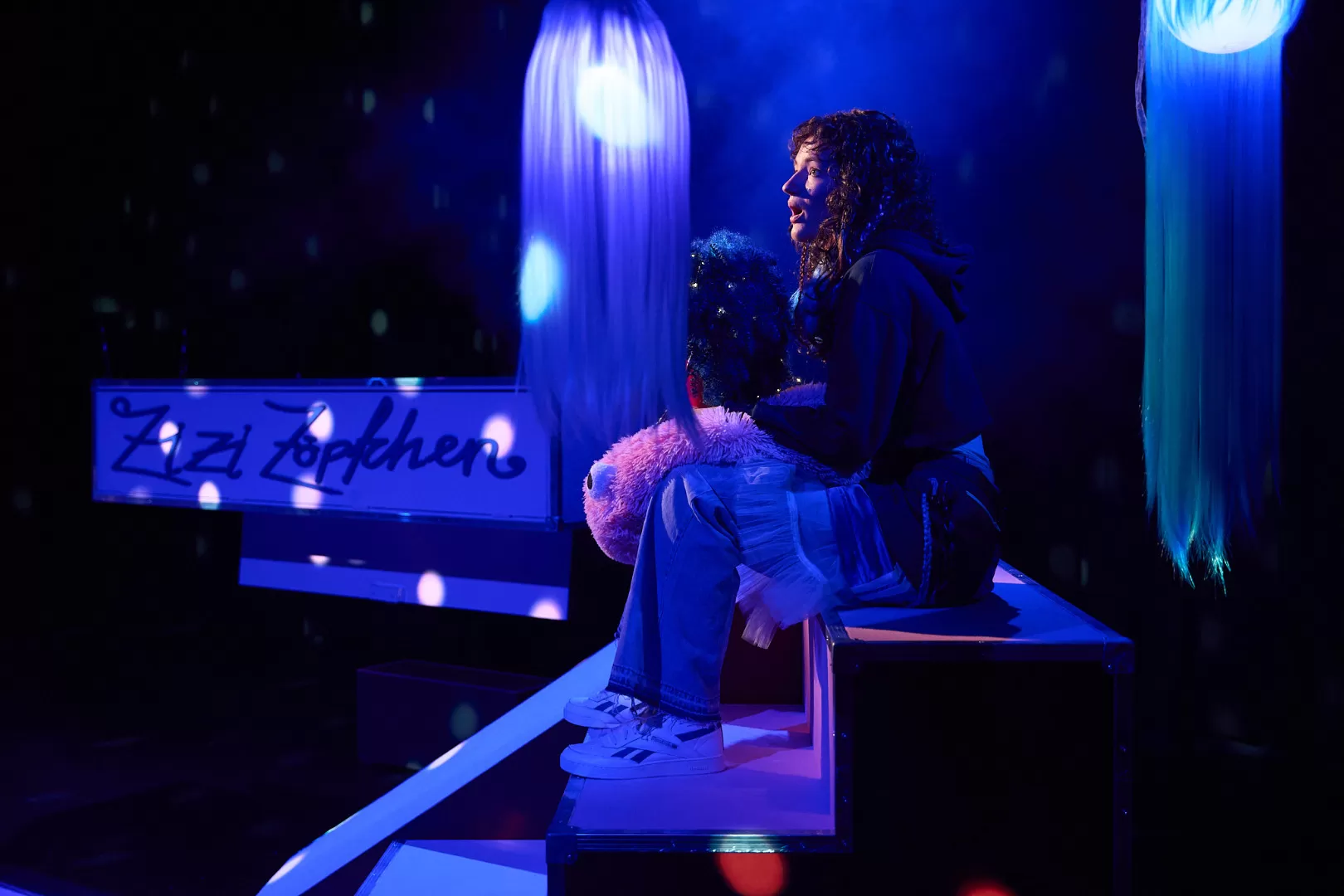 Eine Person mit Locken hält eine Lockenperücke in der Hand und sitzt auf einer Stufe in blauem Licht