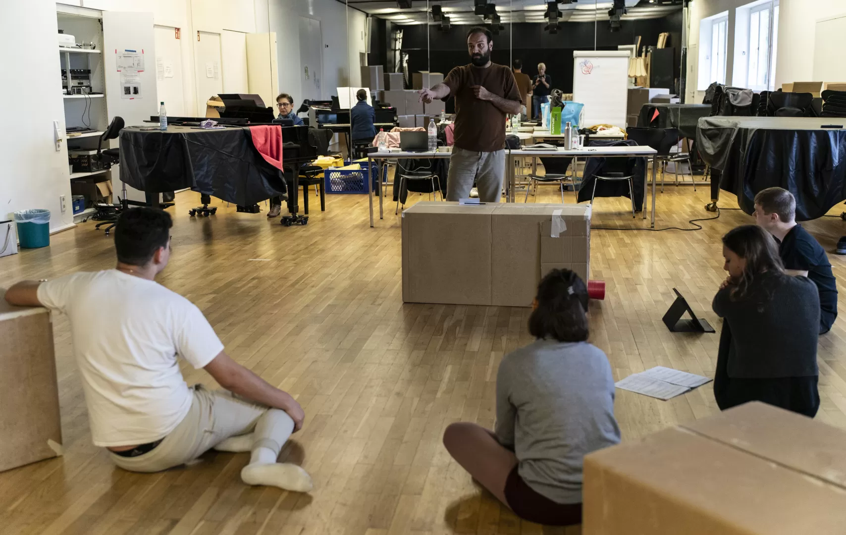 Proben im Opernstudio, ein Mann gibt Regieanweisungen, während die Darstellenden auf dem Boden sitzen