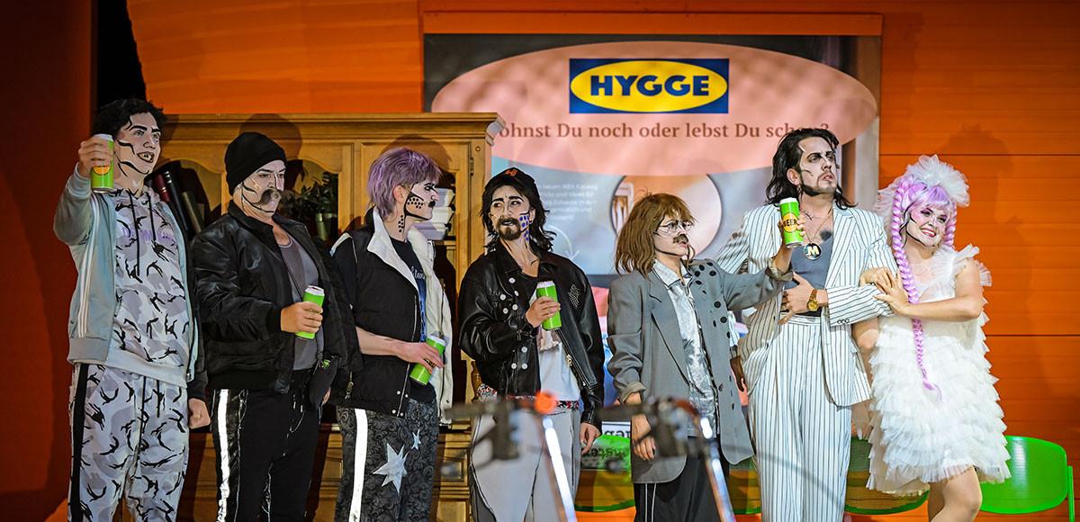 Eine große Personengruppe, alle tragen schwarze, weiße oder graue Klamotten. Sie sind im Pop Art Stil geschminkt. Der Hintergrund ist orange