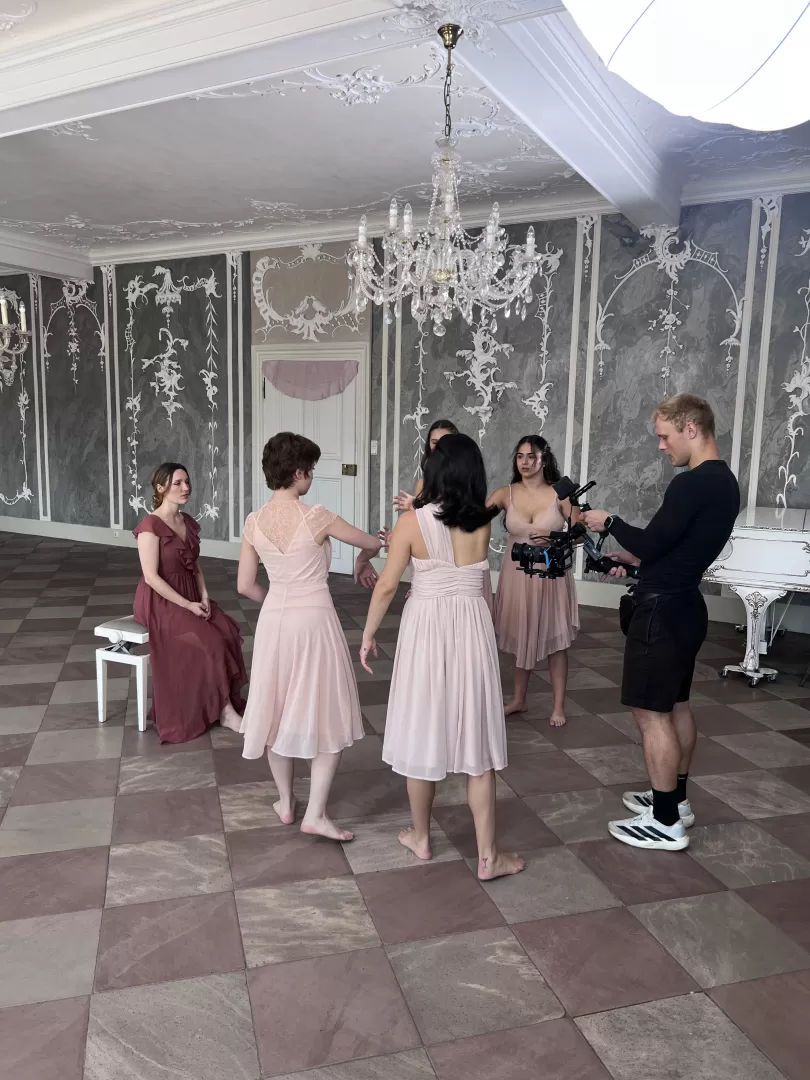 Frauen in rosafarbenen Kleidern werden von einem jungen schwarz gekleideten Mann in einem Schloss gefilmt