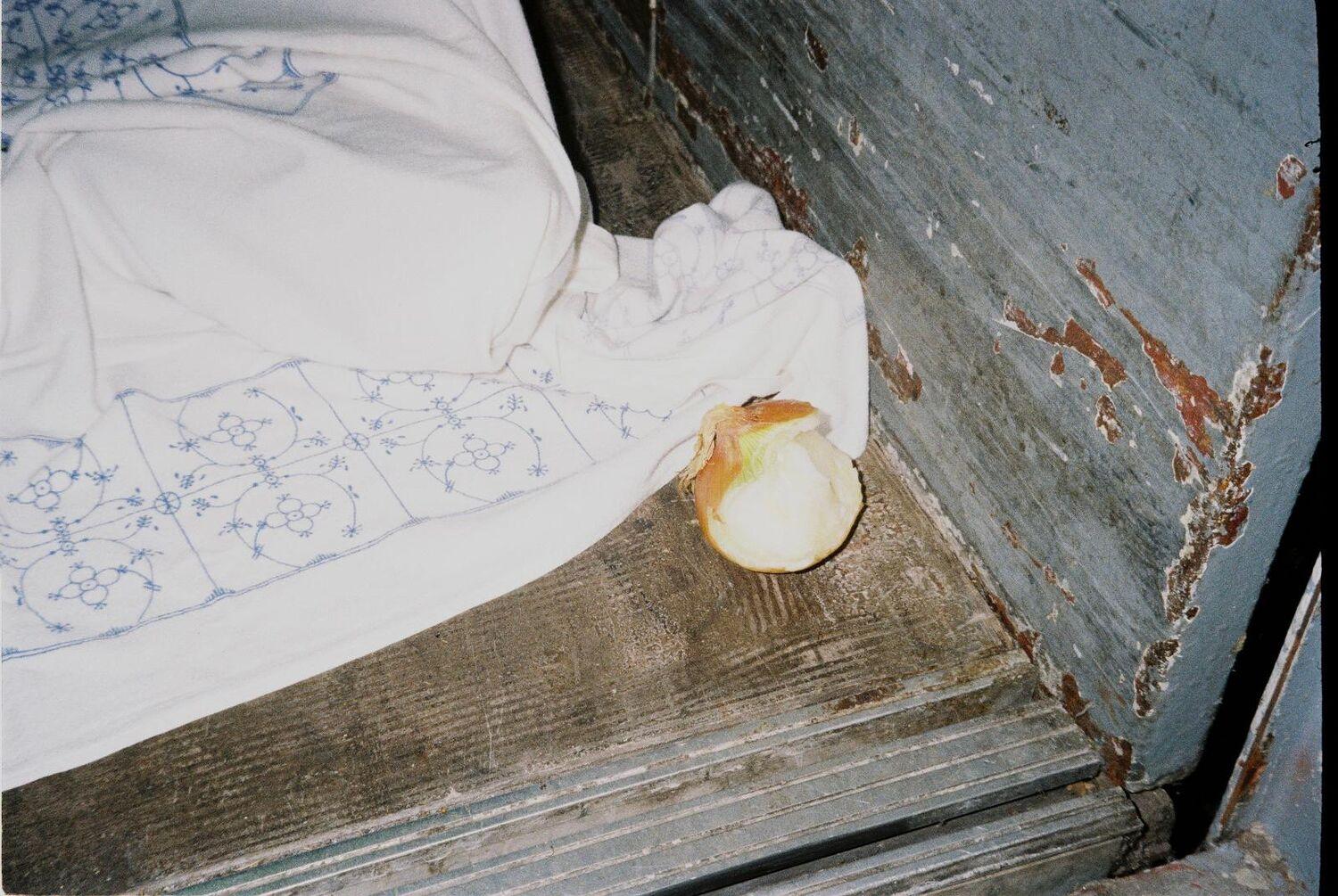 Auf einem alten Holzboden liegt eine blau-weiße Stoffdecke und daneben eine halb geschälte und angebissene Zwiebel.