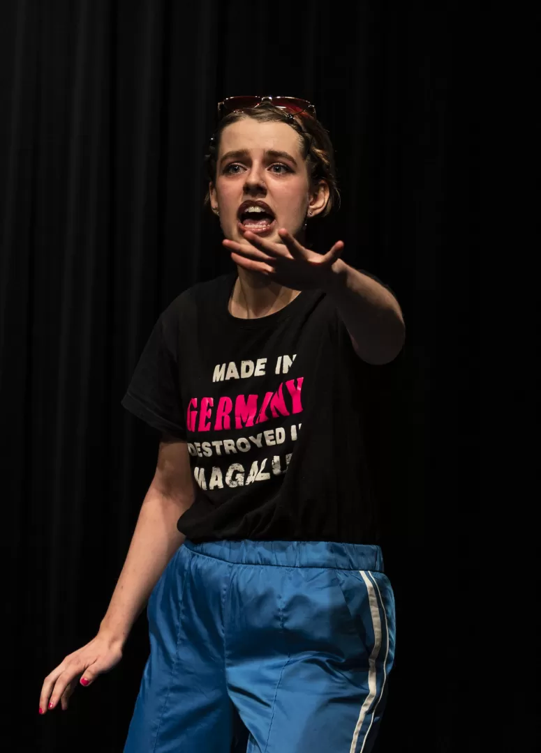 Paula Marie Schulze mit  grell blauer Hose mit weißen Streifen und schwarzem Tshirt mit der Aufschrift “made in Germany, destroyed in Magaluf” in pink. Erstaunter Ausdruck, die rechte Hand ist erhoben. Eine pinke Brille in den Haaren.