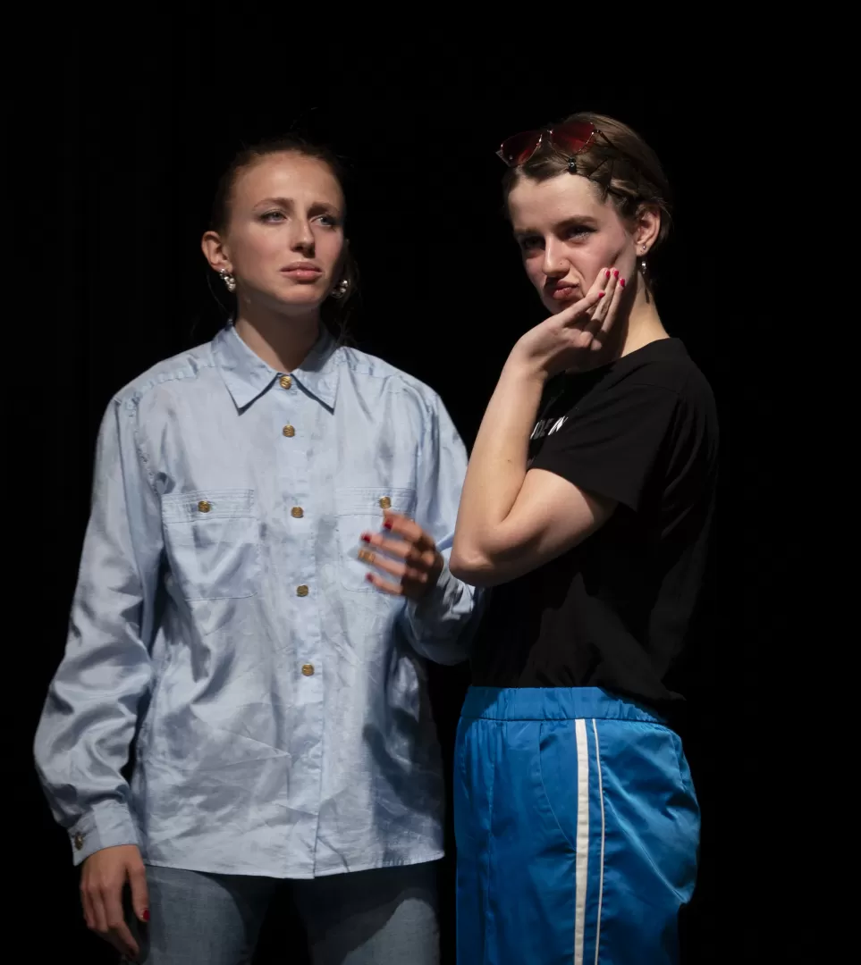 Paula Marie Schulze mit grell blauer Hose  mit weißen Streifen und schwarzem Tshirt mit der Aufschrift “made in Germany, destroyed in Magaluf” in pink. Eine pinke Brille in den Haaren.  Due rechte Hand am Gesicht, drückt die Wangen zu einem Kussmund. Links daneben steht Philine Heyer in einer hellblauen Bluse.