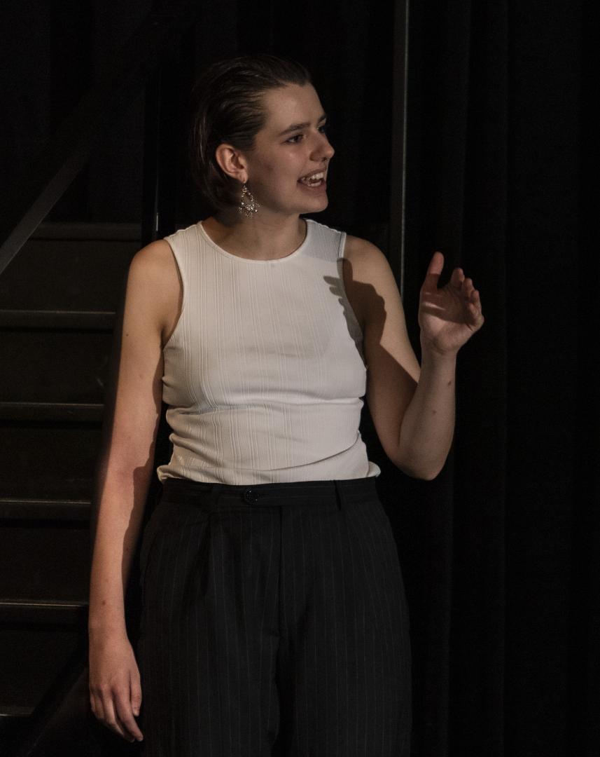 Paula Marie Schulze mit schwarzer Anzugshose und weißem Top. Der linke Arm ist leicht erhoben. Im Hintergrund eine Treppe.