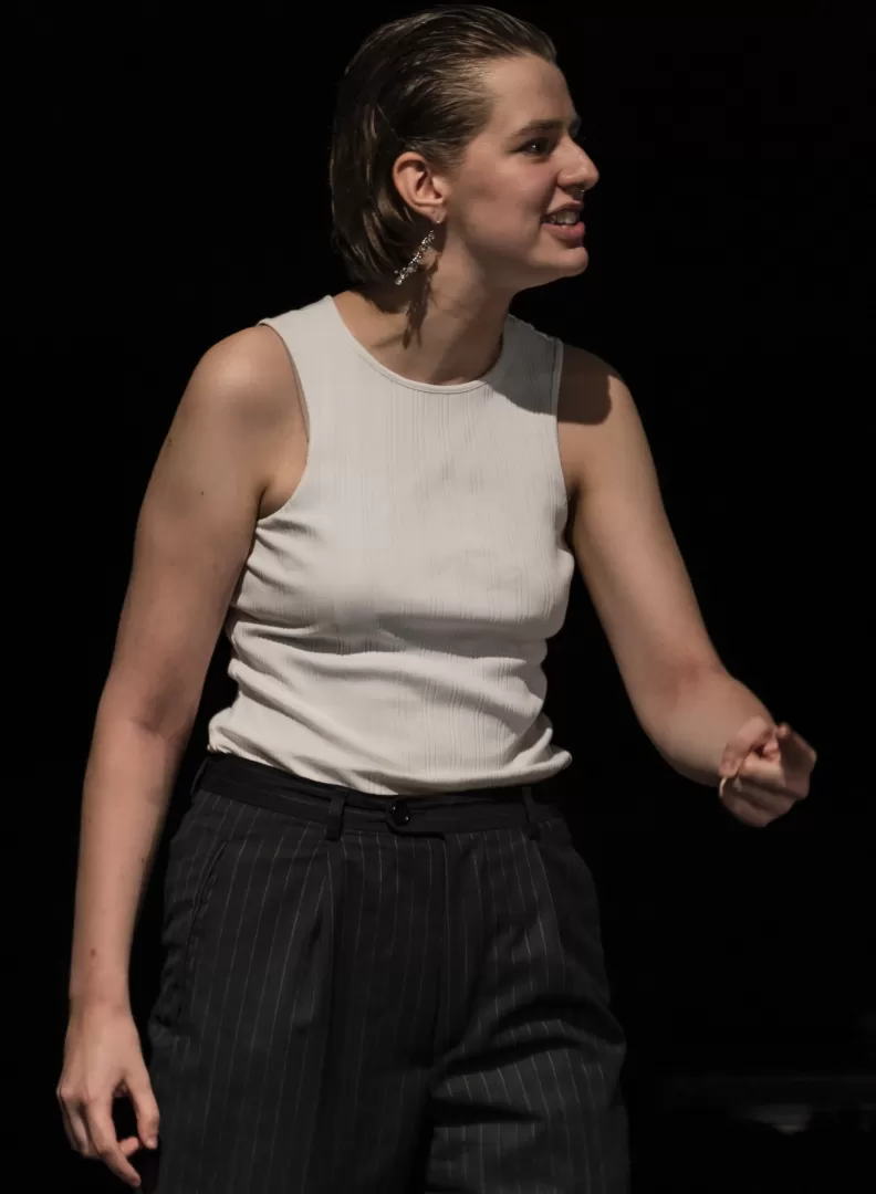 Paula Marie Schulze mit schwarzer Anzugshose und weißem Top. Der linke Arm ist leicht erhoben. Sie zeigt auf etwas, der Ausdruck ist wütend. 