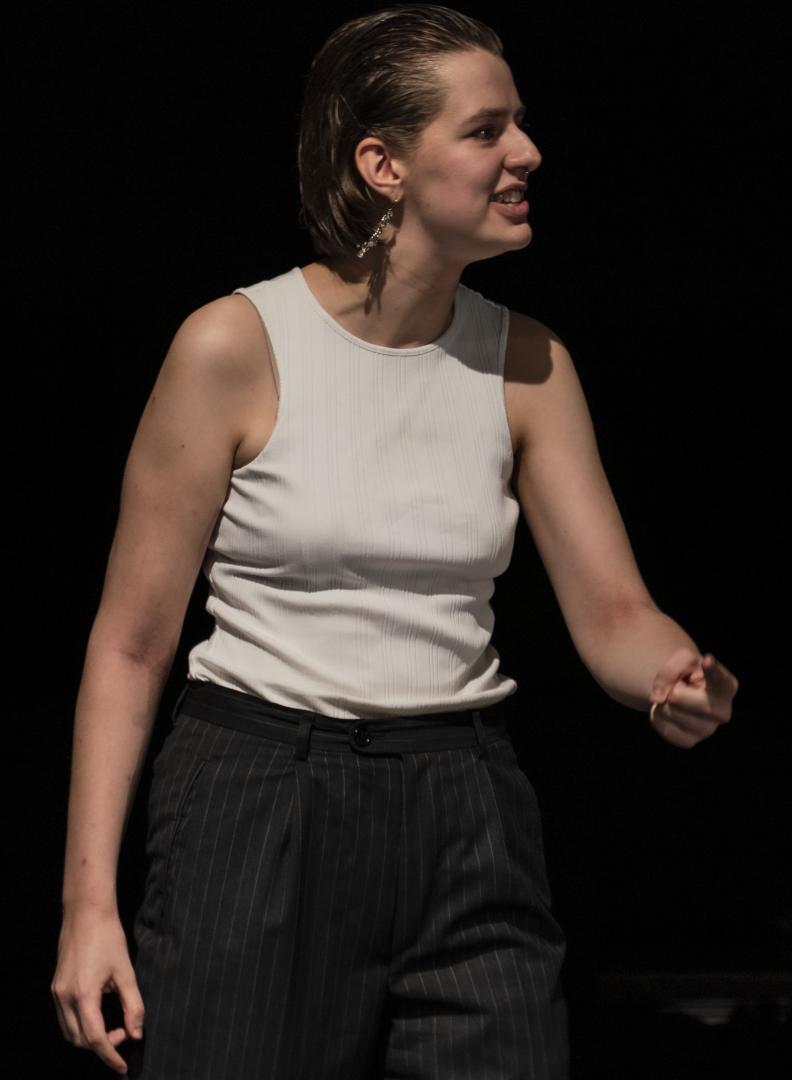 Paula Marie Schulze mit schwarzer Anzugshose und weißem Top. Der linke Arm ist leicht erhoben. Sie zeigt auf etwas, der Ausdruck ist wütend.