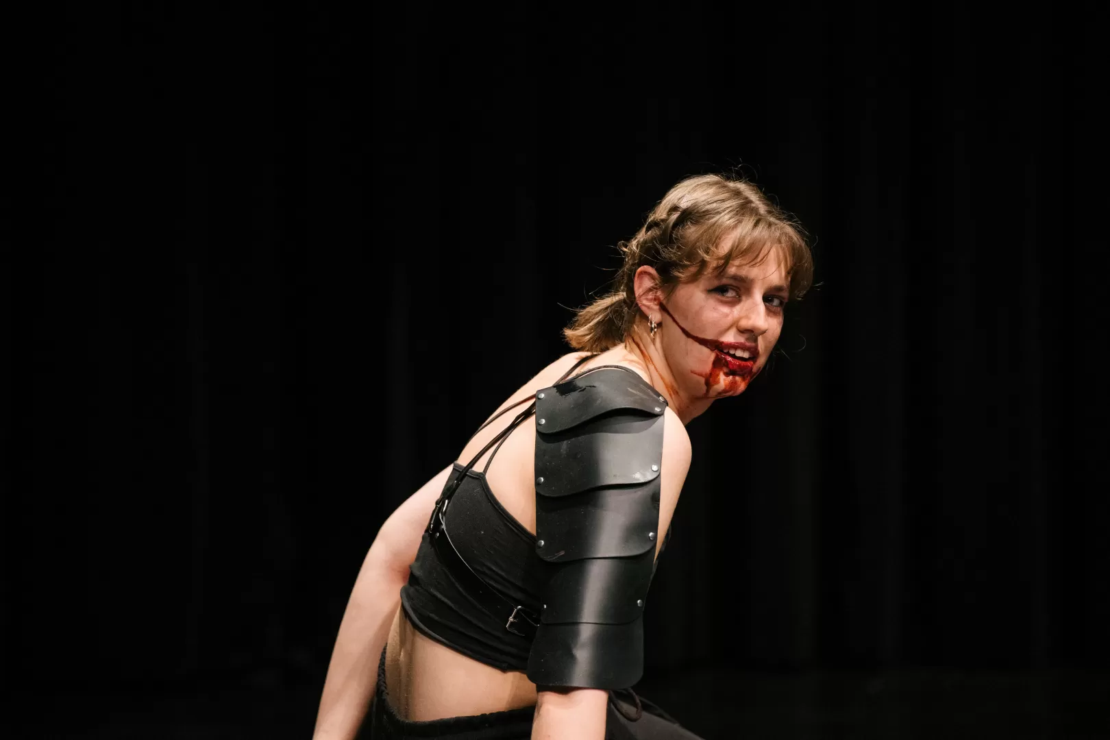 Paula Marie Schulze mit blutverschmiertem Mund, schwarze Kleidung, eine schwarze Lederplatte über der linken Schulter. 