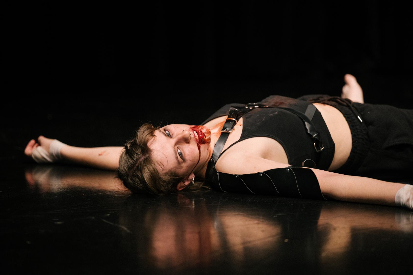 Paula Marie Schulze mit blutverschmiertem Mund, schwarze Kleidung, eine schwarze Lederplatte über der linken Schulter. Sie liegt auf dem Boden, der Kopf ist zur Seite geneigt.