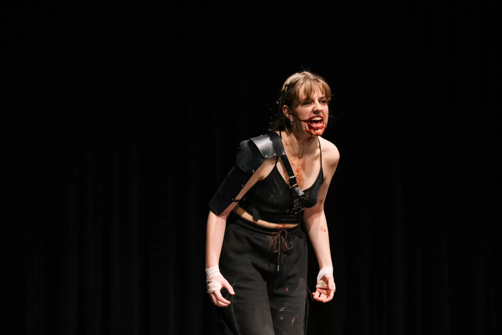 Paula Marie Schulze mit blutverschmiertem Mund, schwarze Kleidung, eine schwarze Lederplatte über der linken Schulter. Der Ausdruck ist wütend, nach vorne gebeugt. 