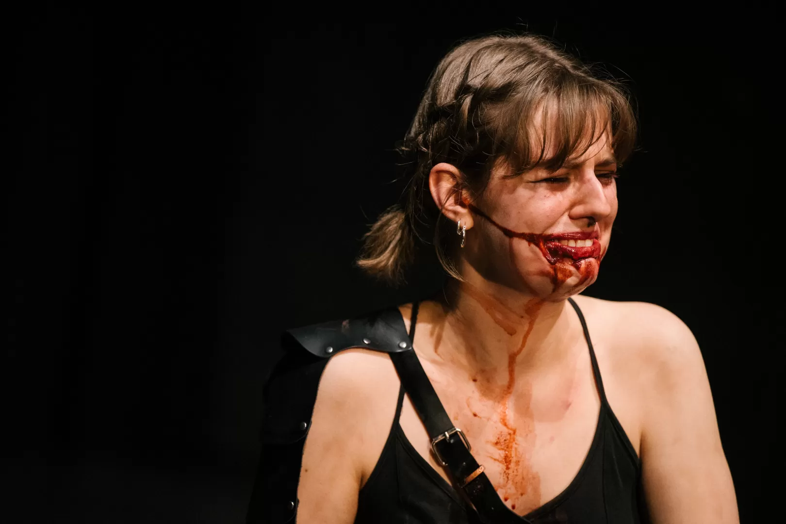 Paula Marie Schulze mit blutverschmiertem Mund, schwarze Kleidung, eine schwarze Lederplatte über der linken Schulter. Der Ausdruck ist trauernd. 