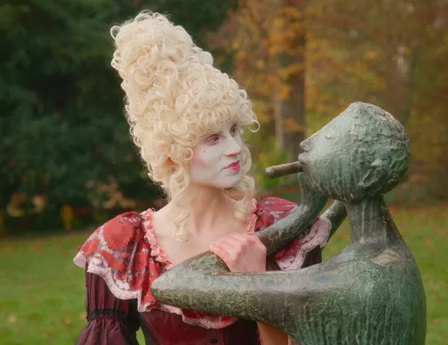 EIne Frau mit barocken Kleidern und Perücke steht neben einer Büste