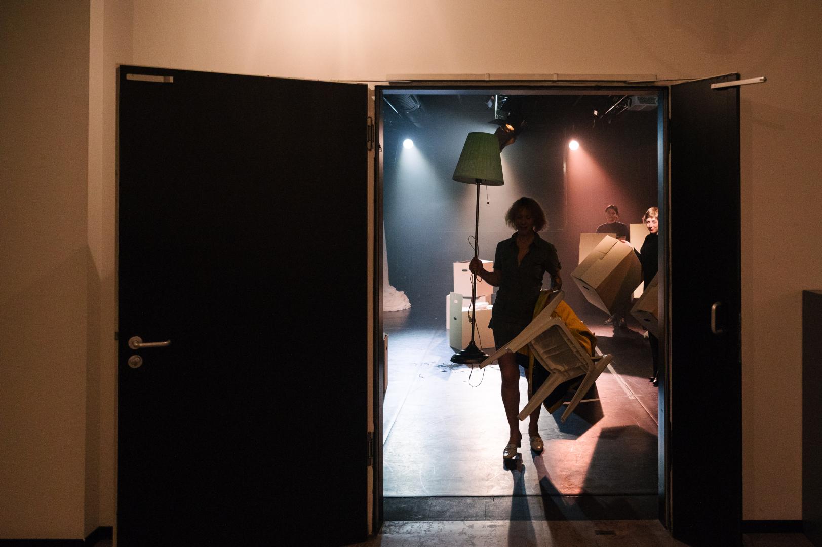 Eine Person mit Stehlampe und Umzugskarton in der Hand kommt aus einem Theatersaal.