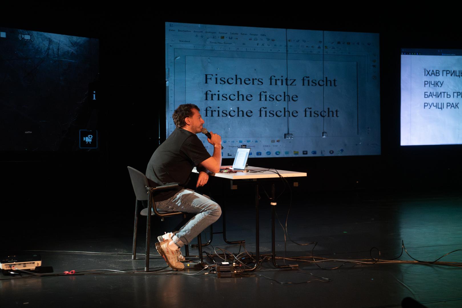 Person mit Handmikrofon sitzt an einem Tisch vor einer Videoprojektion.