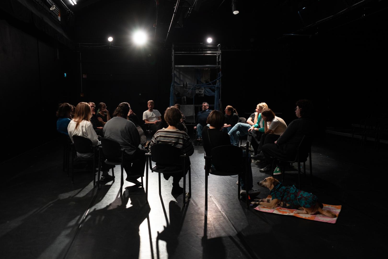 Personen sitzen in einem Stuhlkreis auf einer Bühnenfläche.