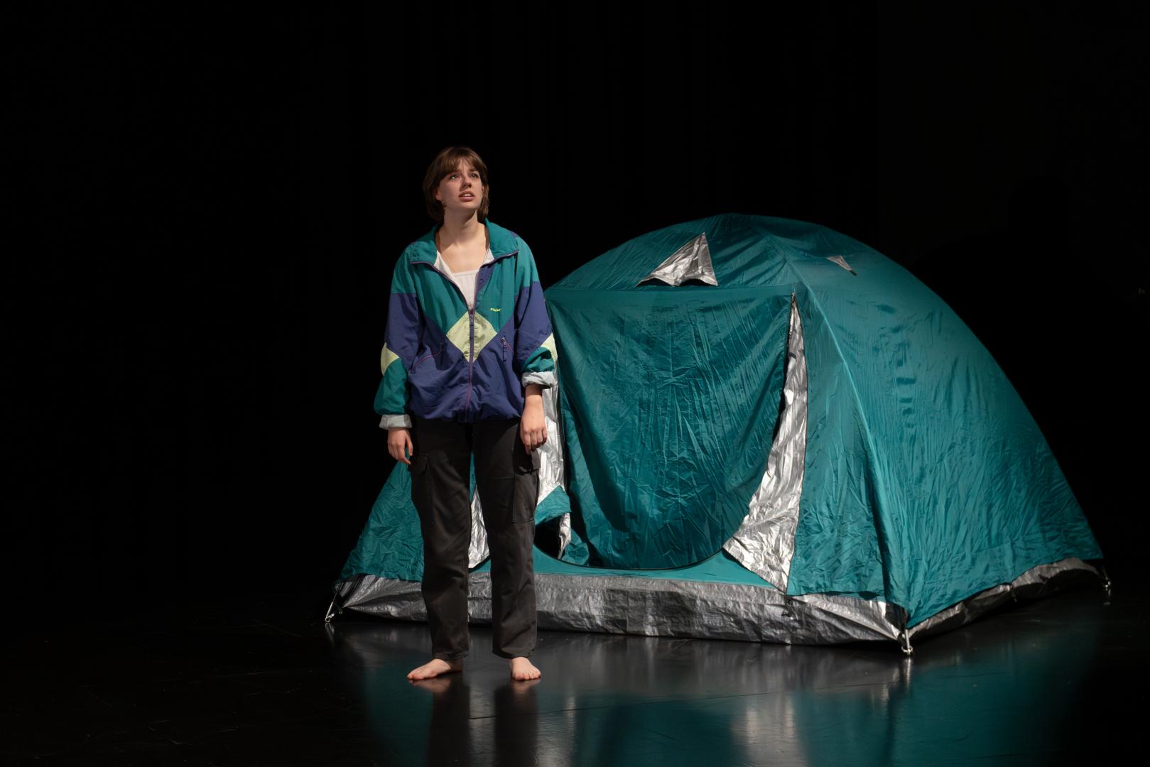 Paula Marie Schulze, grün-blaue und lila Jacke, schwarz-graue Cargohose, auf dem schwarzen Bühnenboden stehend. Rechts dahinter in der Bühnenecke ein grün-blaues Zelt.