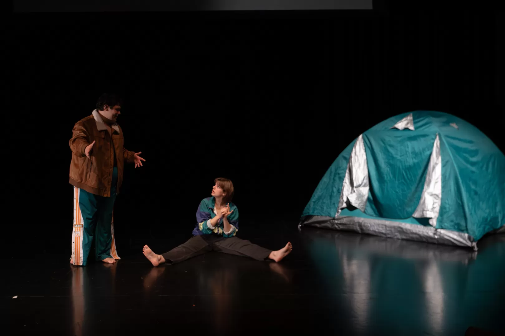 Paula Marie Schulze, grün-blaue und lila Jacke, schwarz-graue Cargohose,  auf dem schwarzen Bühnenboden sitzend, die Hände hoch haltend, mit den Fingern zählend. Links daneben Kevin Krougliak in brauner Jacke stehend. Rechts daneben in der Bühnenecke ein grün-blaues Zelt.
