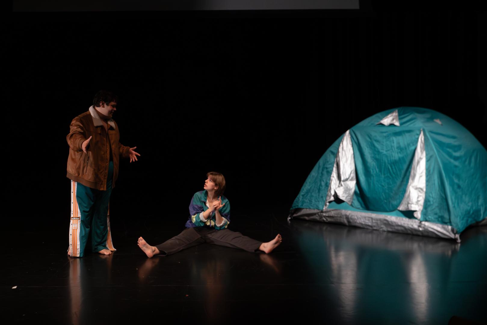 Paula Marie Schulze, grün-blaue und lila Jacke, schwarz-graue Cargohose, auf dem schwarzen Bühnenboden sitzend, die Hände hoch haltend, mit den Fingern zählend. Links daneben Kevin Krougliak in brauner Jacke stehend. Rechts daneben in der Bühnenecke ein grün-blaues Zelt.