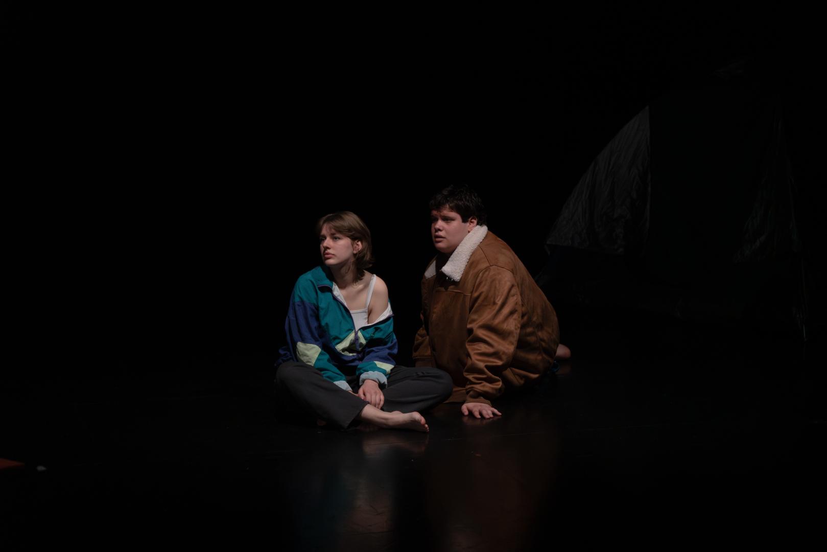 Paula Marie Schulze, grün-blaue und lila Jacke, schwarz-graue Cargohose, auf dem schwarzen Bühnenboden sitzend. Rechts dahinter Kevin Krougliak in brauner Jacke sitzend. Rechts daneben in der Bühnenecke ein grün-blaues Zelt.