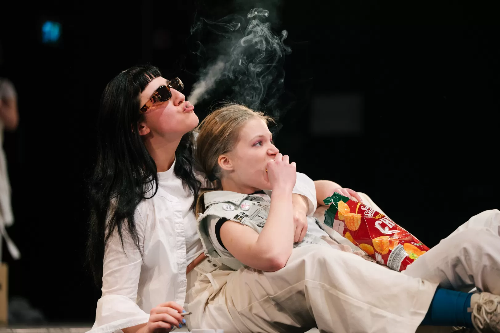 Eine Frau liegt chipsessend im Schoß einer anderen Frau, die Zigarettenrauch auspustet.