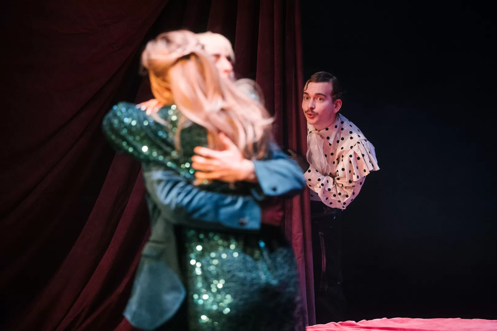 Ein Mann schaut hinter einem Vorhang hervor und sieht, wie sich zwei Personen umarmen.