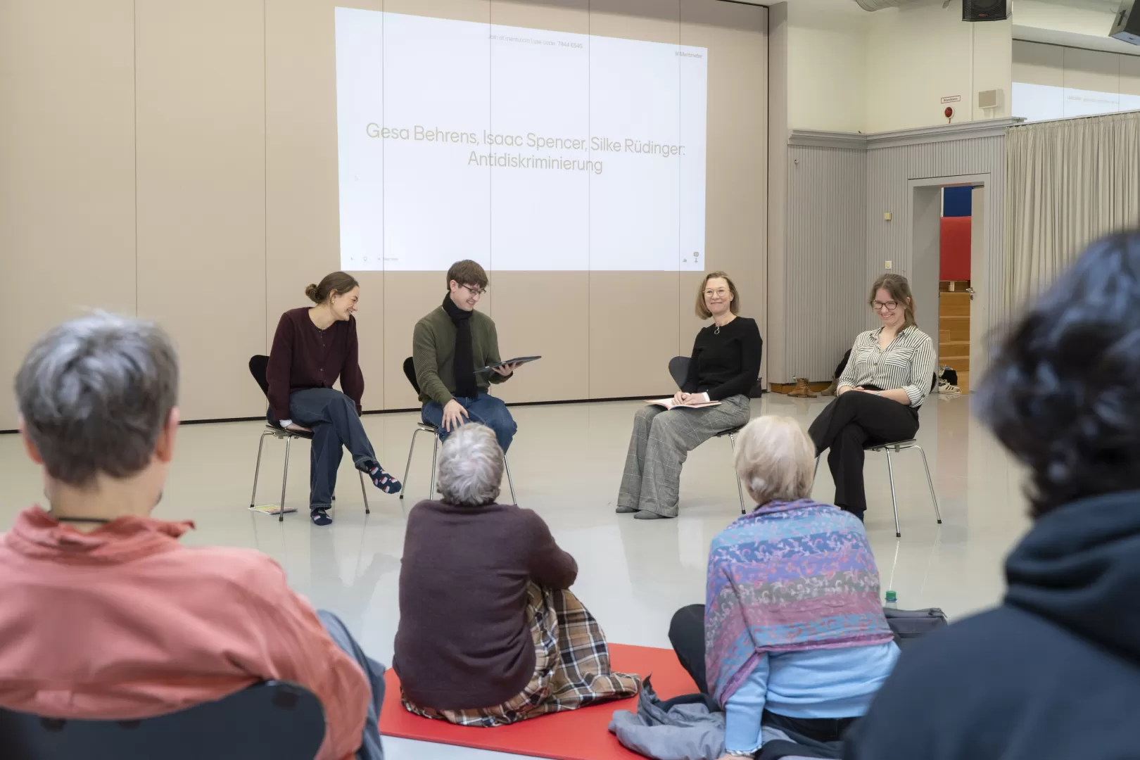 Vier Personen sitzen vor einer Gruppe Zuhörer*innen