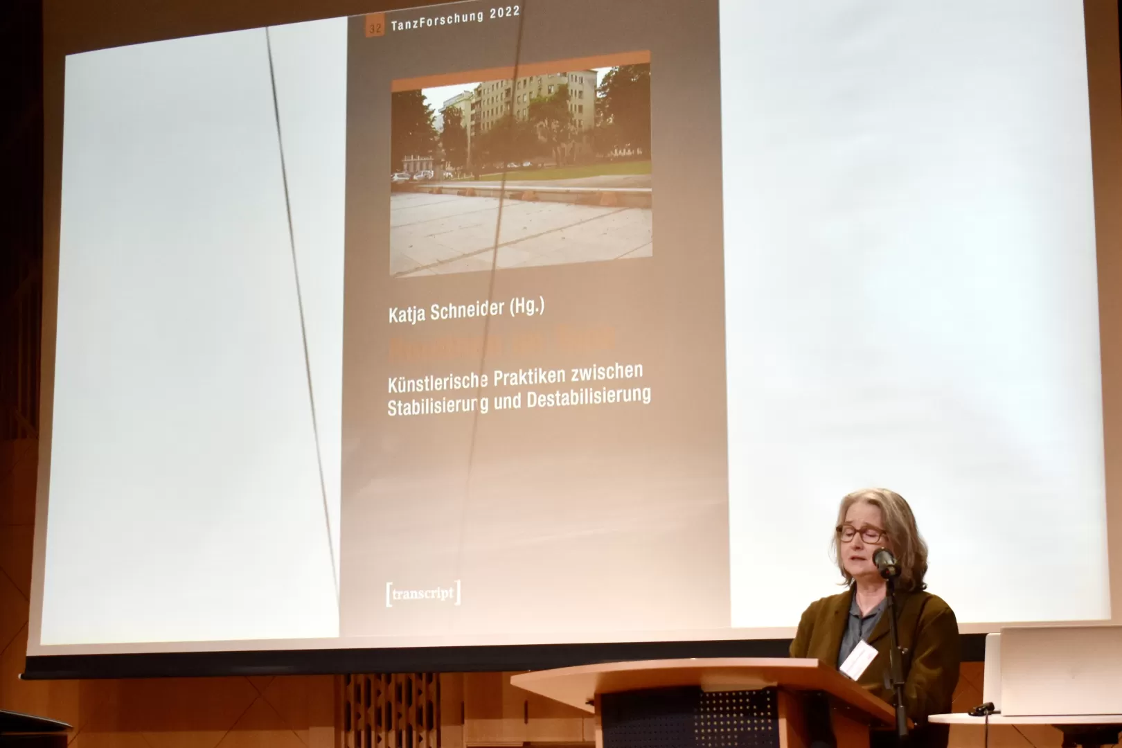 Katja Schneider steht am Redepult im Kleinen Saal der HfMDK, hinter ihr auf der Präsentationsleinwand ist das Cover des von ihr herausgegebenen Buchs "Routinen im Tanz" zu sehen. 