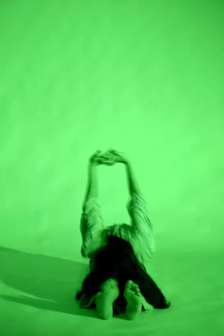 A person sitting on the floor, bending to her bare feet with hands locked behind her. Green light on her and in the background 
