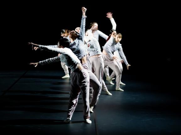 Eine Gruppe Tänzerinnen in Performance, hell gekleidet vor dunklem Bühnenhintergrund. 