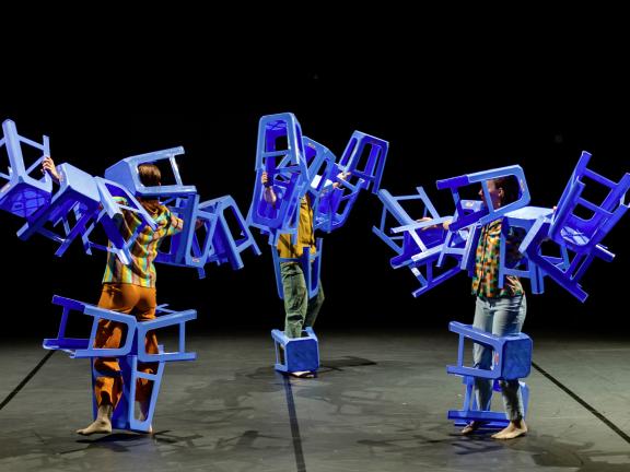 Szene beim Tanzmarathon 2021, drei Personen tragen viele blaue Hocker an ihren Körpern und bewegen sich auf der Bühne.
