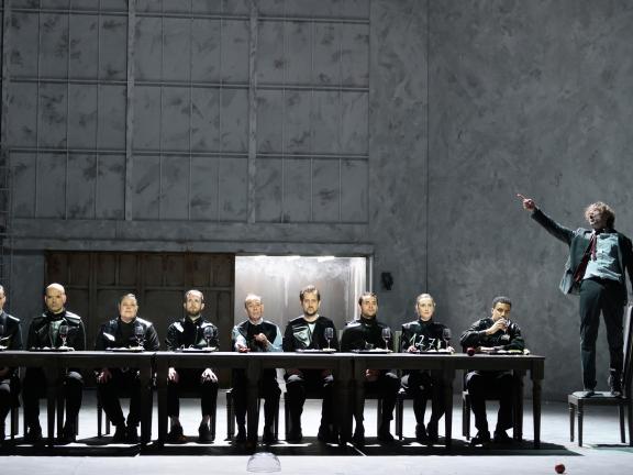 An einer langen Tafel sitzen neun schwarz gekleidete Personen, auf einem Stuhl am Kopfende steht ein Schauspieler.