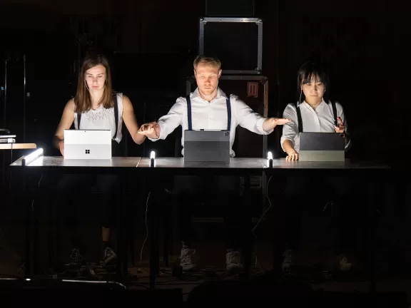 Drei Performer*innen im dunklen Raum, vor ihnen Laptops auf dem Tisch.