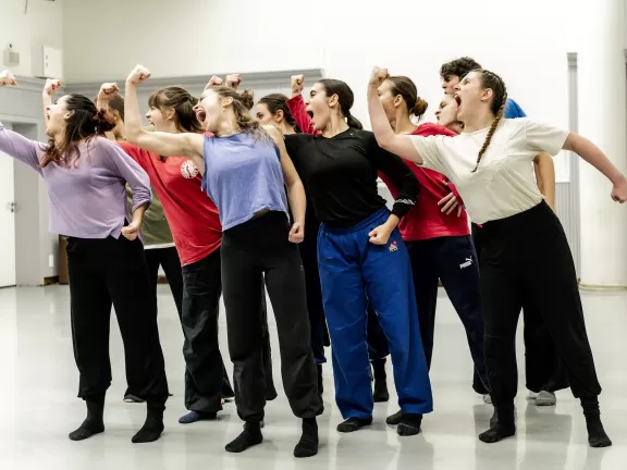 Eine Gruppe von Tänzer*innen, die sich nach links lehnen und die rechte Hand erhoben mit einem 45 Grad Winkel Knick schreien.
