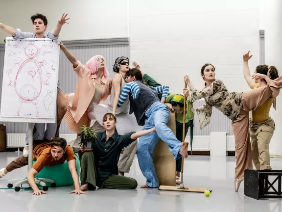 Eine Gruppe von Tänzer*innen, die ineinander verhakt dargestellt sind mit Bühnen Props wie einem Stuhl oder einem Plakat.