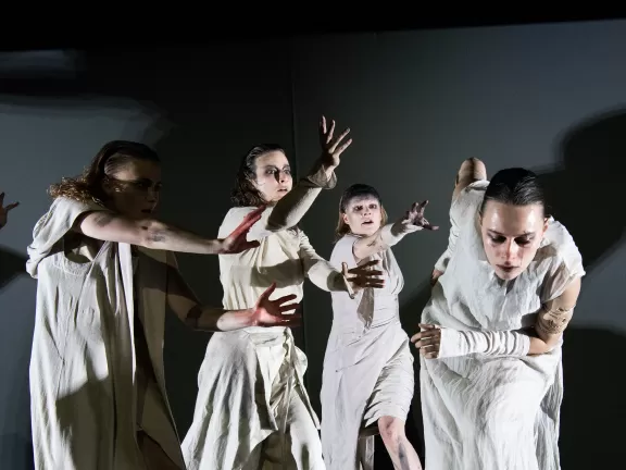 Vier Performer*innen in weißen Toga-Gewändern bewegen sich schnell und dynamisch auf der Bühne