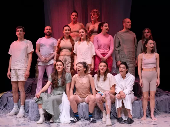 Große Gruppe Darsteller*innen in sommerlicher Kleidung in einer Theaterszene vor schwarzer Wand.