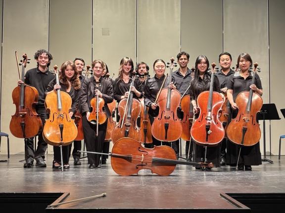 California Cello Ensemble & Studierende der HfMDK Die Mitglieder eines größeren Cello-Ensembles stehen mit ihren Instrumenten auf einer Bühne, Gruppenfoto.