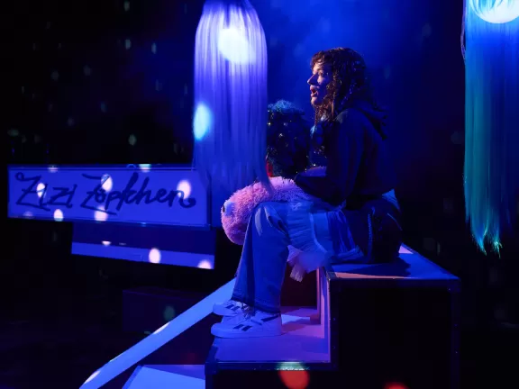 Eine Person mit Locken hält eine Lockenperücke in der Hand und sitzt auf einer Stufe in blauem Licht
