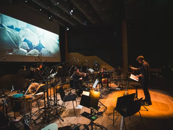 Ein Dirigent dirigiert ein Ensemble live zu einem Film, der auf der Leinwand gezeigt wird.