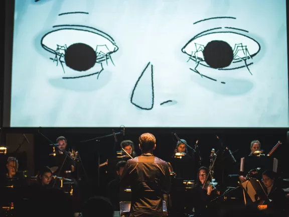 Dirigient vor einem Ensemble, im Hintergrund eine Leinwand auf der ein Stummfilm