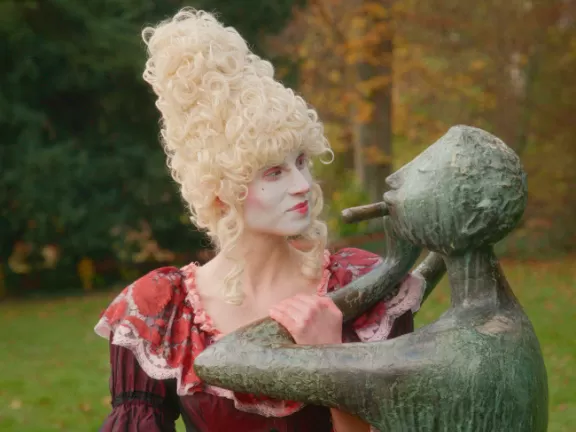 EIne Frau mit barocken Kleidern und Perücke steht neben einer Büste