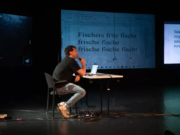 Mann mit Handmikrofon sitzt an einem Tisch vor einem Laptop.