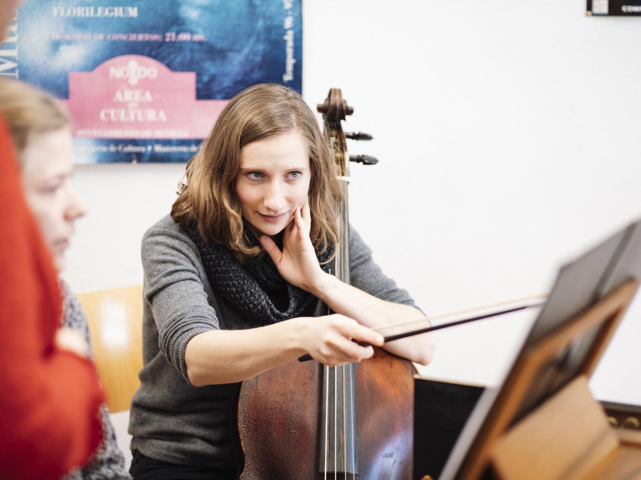 Eine junge Frau mit Cello zeigt mit dem Streicherbogen auf Noten. 