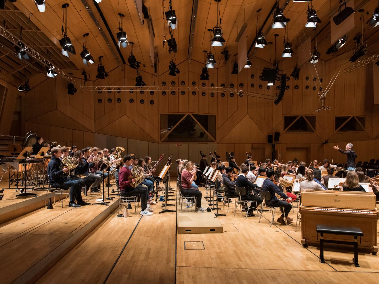 Seitliche Ansicht eines Orchesters auf der Bühne, rechts im Bild der Dirigent.