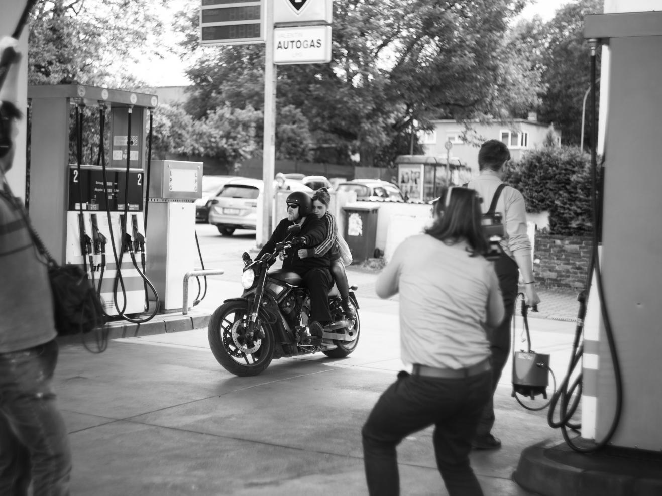 Zwei Personen fahren auf einem Motorrad in eine Tankstelle und werden dabei gefilmt.