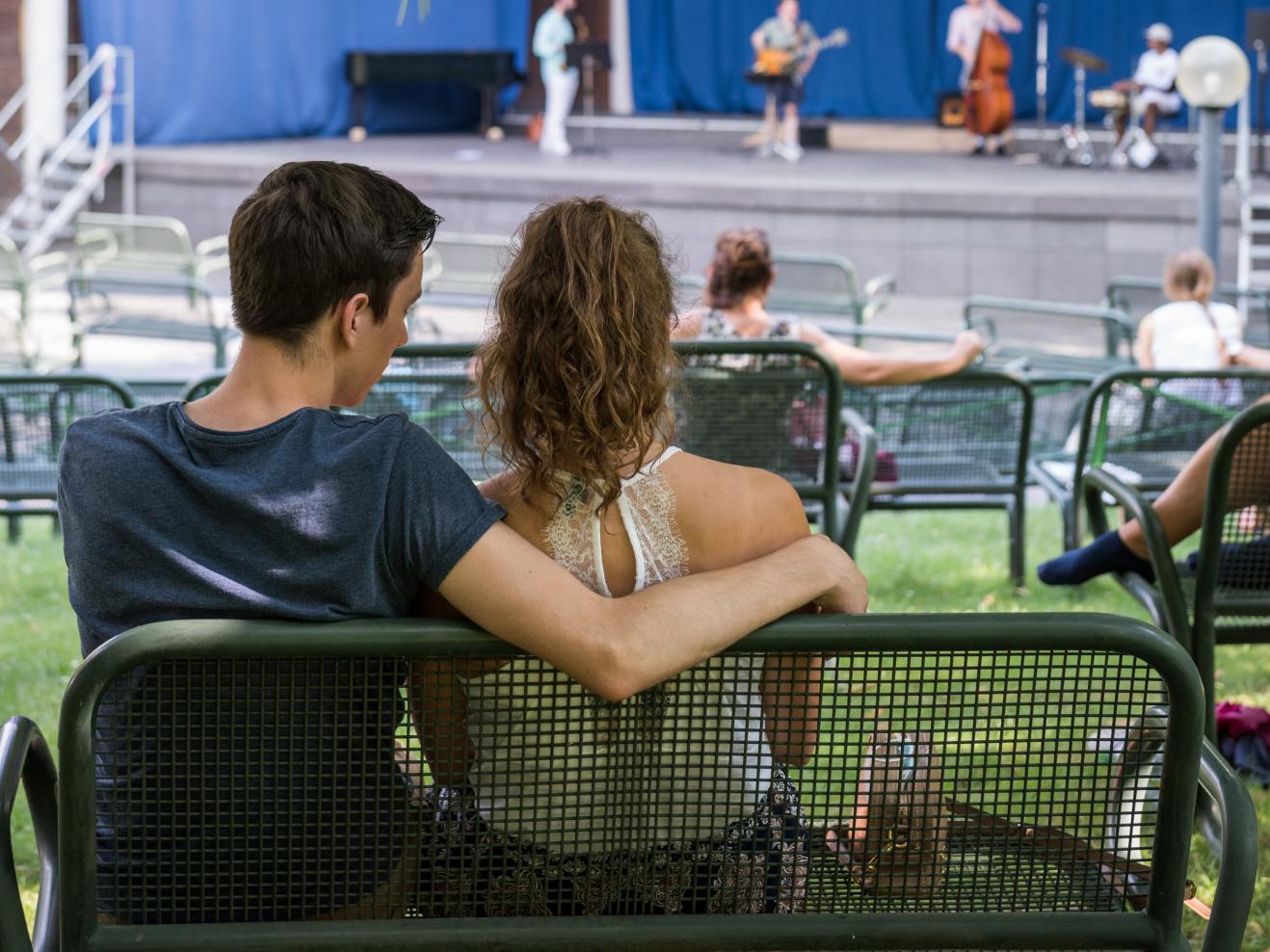 Eine junge Frau sitzt im Arm eines jungen Mannes auf einer Bank. Auf der Bühne spielt ein Jazzquartett